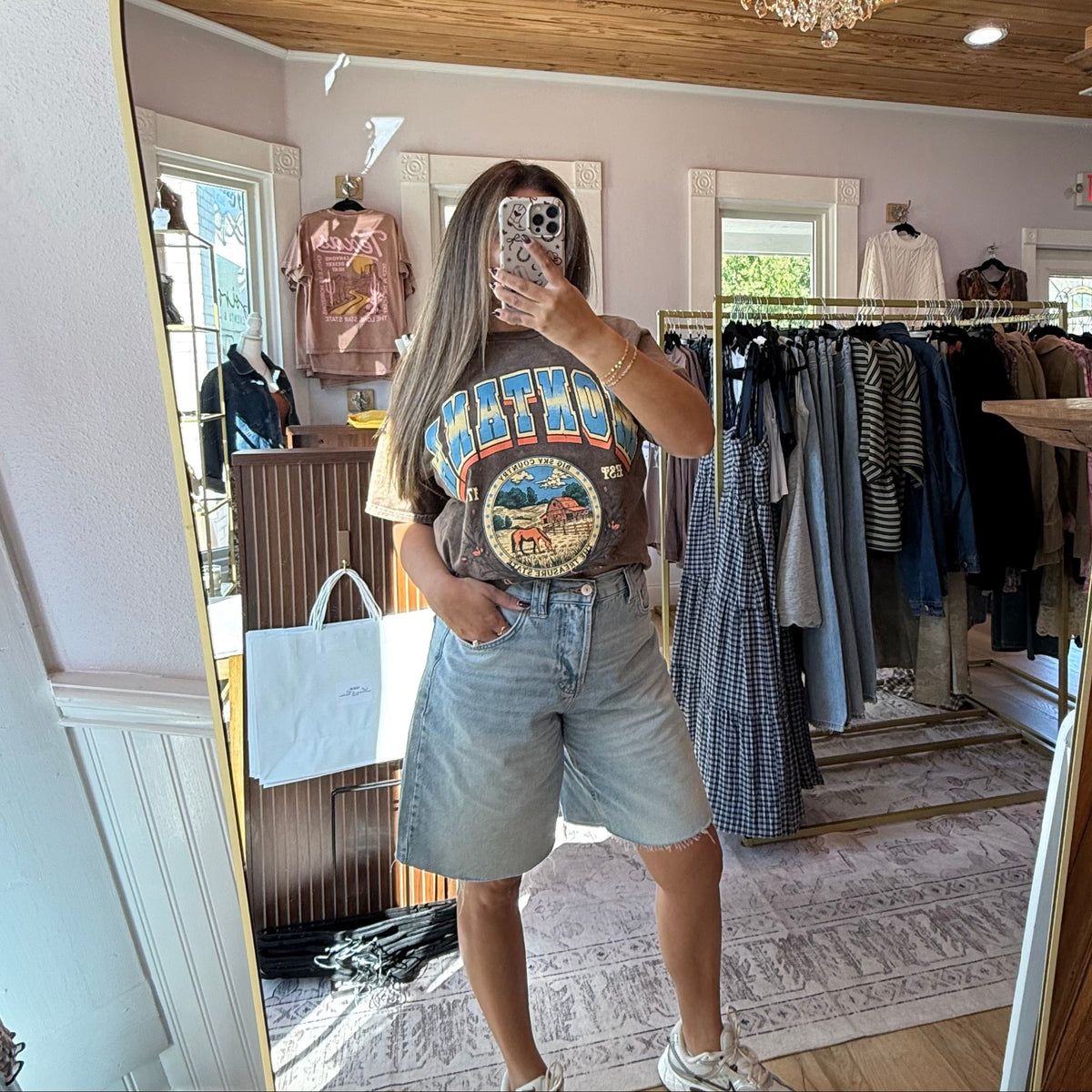 A woman takes a mirror selfie in a boutique while wearing a brown Montana graphic t-shirt and light blue denim shorts. The boutique interior features a wooden ceiling, a crystal chandelier, and clothing racks in the background.