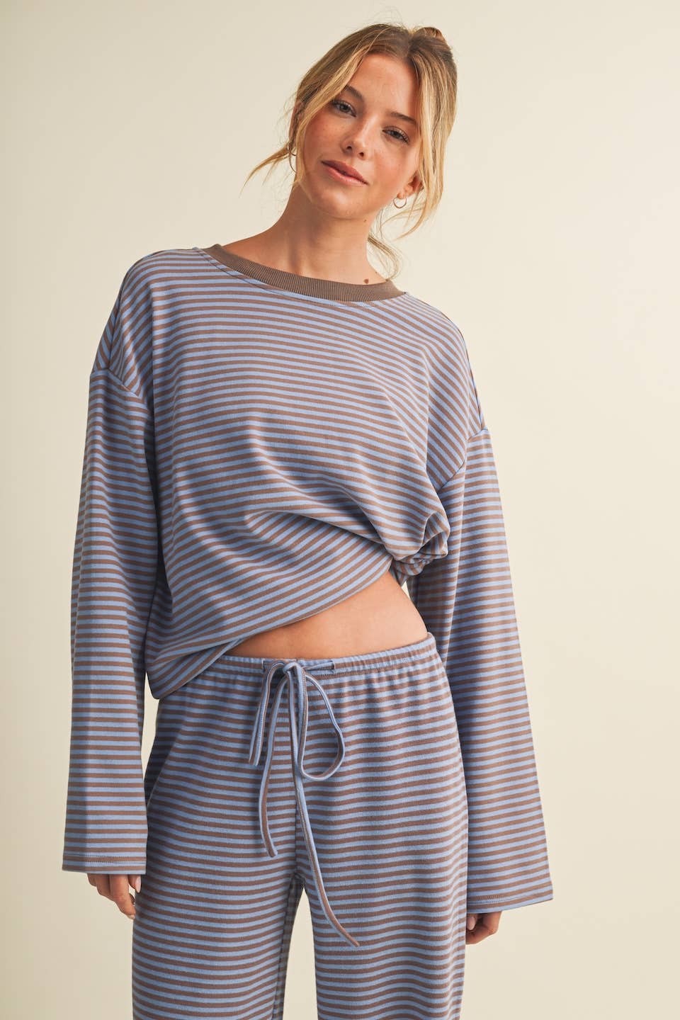 A woman modeling a matching two-piece loungewear set featuring a long-sleeve cropped top and drawstring pants, both in a muted blue-gray and taupe striped pattern, against a solid light beige background.
