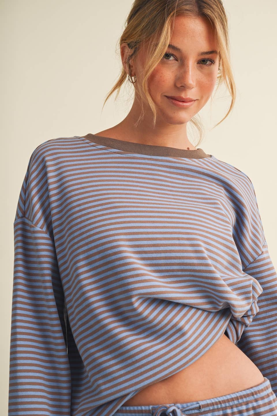A woman wearing a long-sleeve cropped top with thin horizontal blue and beige stripes, styled with matching striped drawstring pants against a plain light beige background.