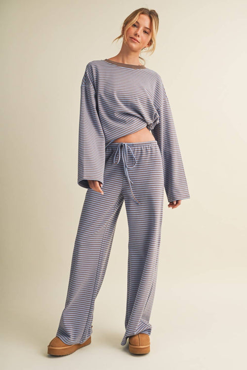 A woman modeling a matching two-piece loungewear set with a long-sleeve cropped top and wide-leg pants, both featuring thin horizontal blue and white stripes, against a solid beige studio background.