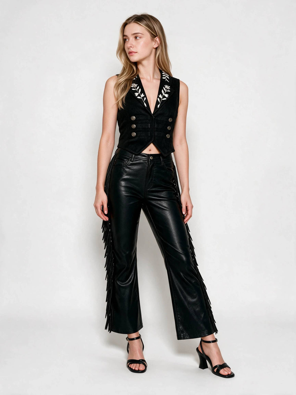 A woman modeling a black sleeveless vest with white embroidered detailing and gold buttons, paired with black fringe pants and black heeled sandals, against a light gray studio background.