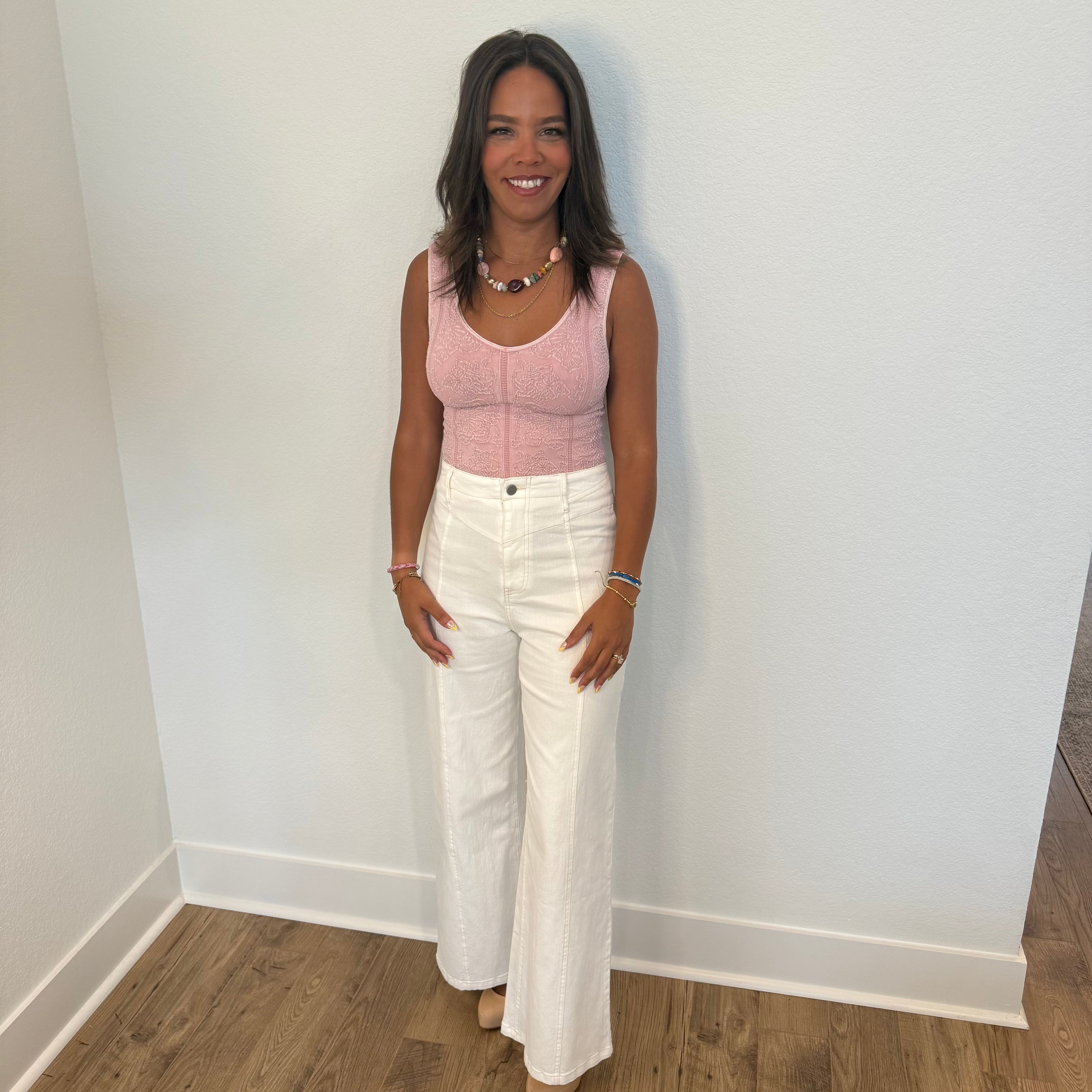 Woman wearing a pink sleeveless top and white pants standing against a plain wall.